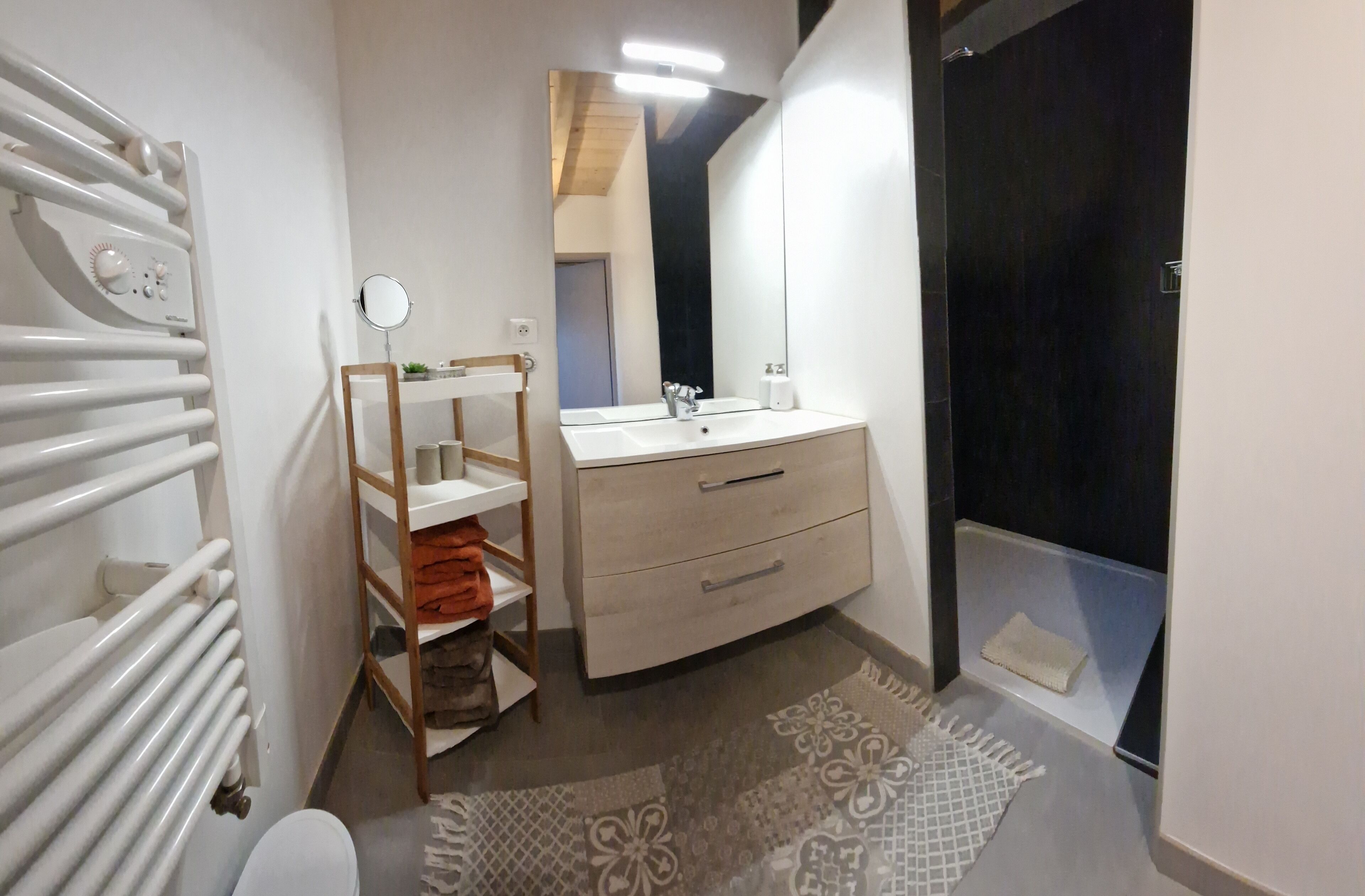 Une salle de bain moderne avec un radiateur blanc, une étagère en bois où sont posées des serviettes, un lavabo blanc avec un miroir et une porte sombre.