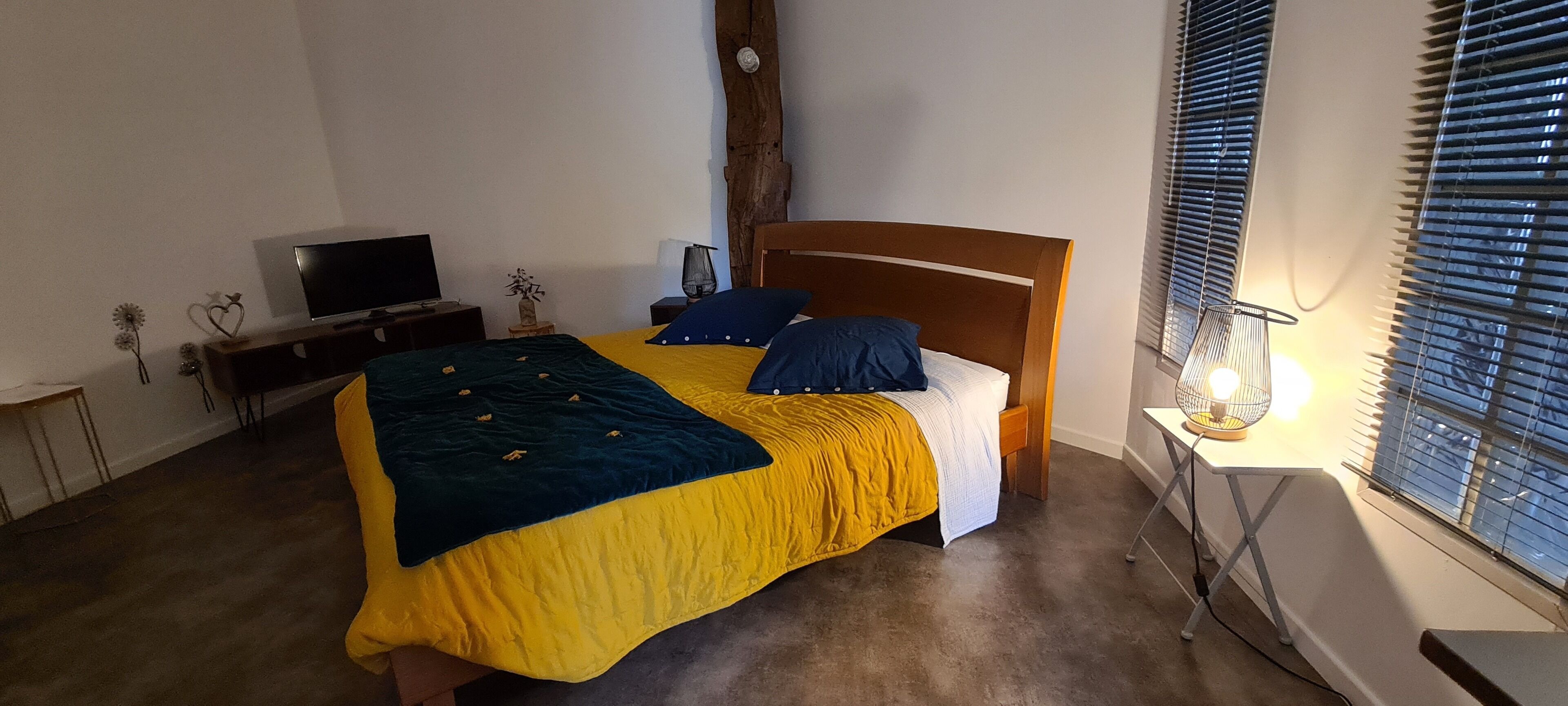 Een slaapkamer met een bed, een televisie, een lamp en een raam met jaloezieën.