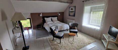 Un dormitorio con cama, una silla, un televisor y un ventanal con cortinas.