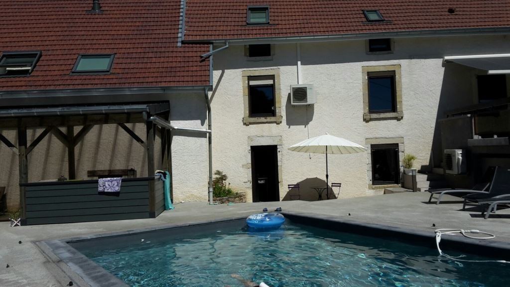 Une piscine avec un espace de détente, un patio couvert et un bâtiment surmonté d’un toit en tuiles rouges.