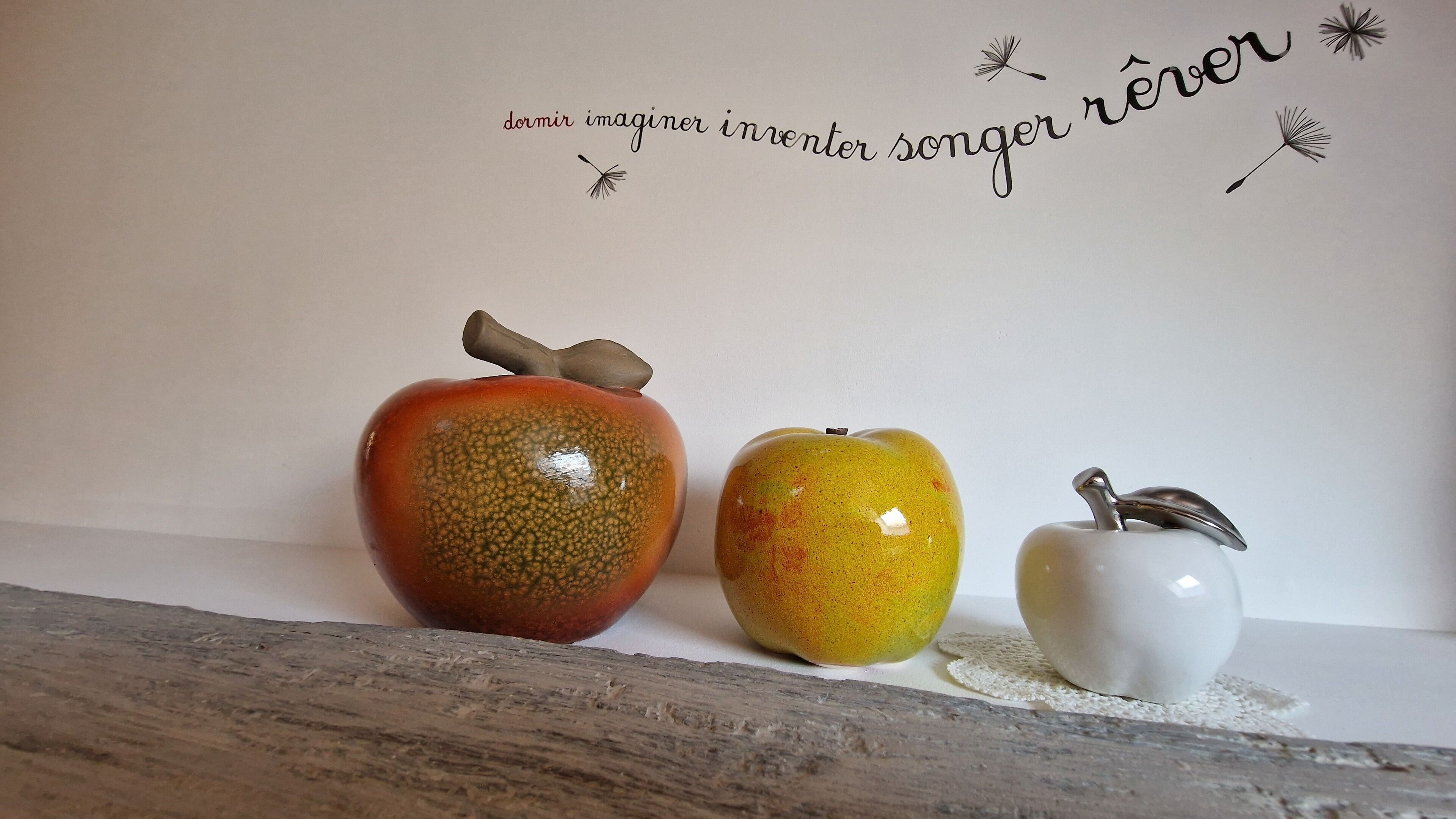 Three ceramic apples with different colors and textures on a wooden surface against a white wall with decorative text.