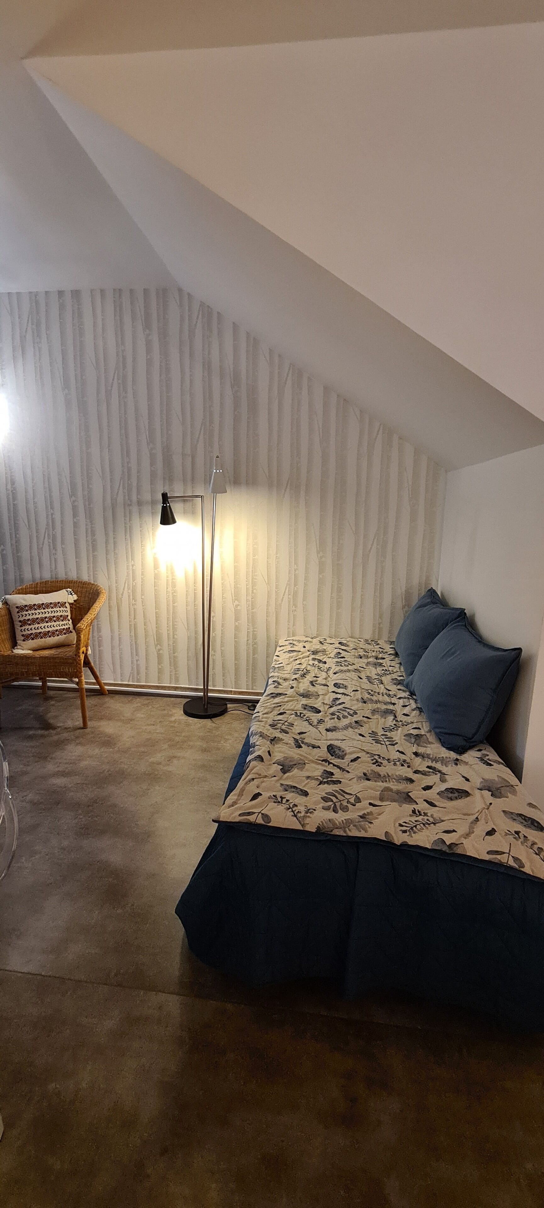Quarto com teto inclinado, cama com colcha florida, duas almofadas, luminária de chão e uma cadeira de vime.