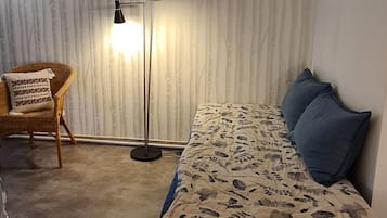 Quarto com teto inclinado, cama com colcha florida, duas almofadas, luminária de chão e uma cadeira de vime.