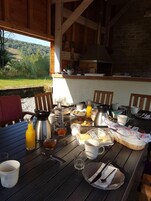 Uma mesa de jantar de madeira com itens de café da manhã, uma cafeteira e vista para uma paisagem arborizada.