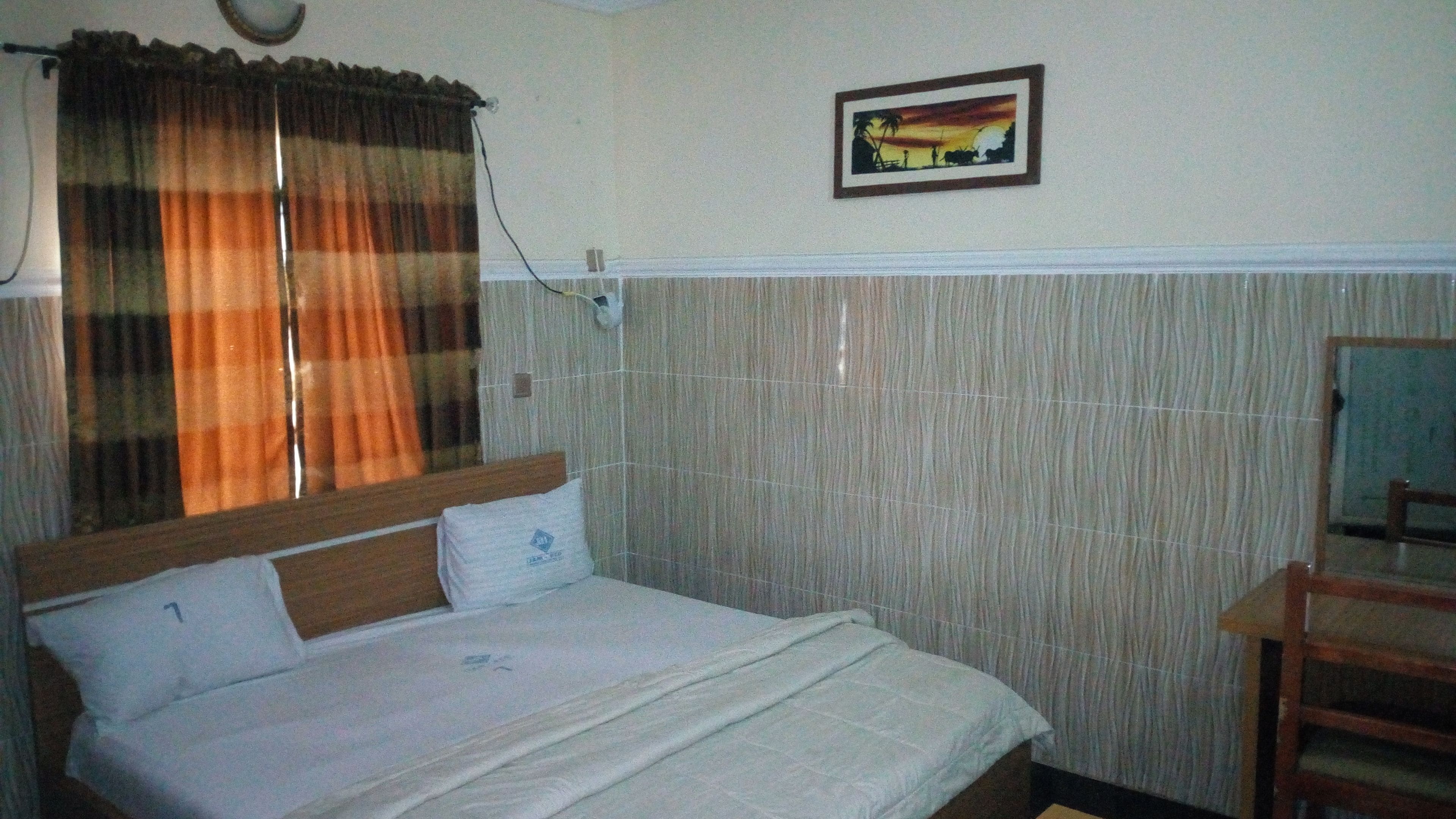 A single-bed room with a wooden headboard, a bedside table, a chair, and a painting on the wall.