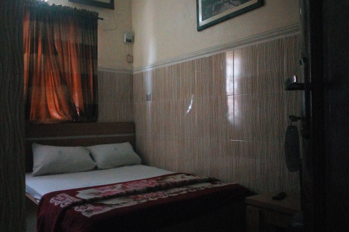 Ein kleines, schwach beleuchtetes Schlafzimmer mit einem Einzelbett, einem Holz-Kopfstück und einem zur Seite gezogenen Vorhang.