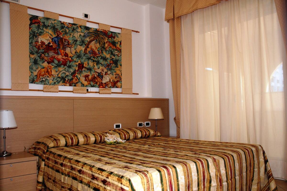 Une chambre d’hôtel avec un grand lit, une table de chevet, une lampe, un téléviseur fixé au mur et une grande tapisserie.