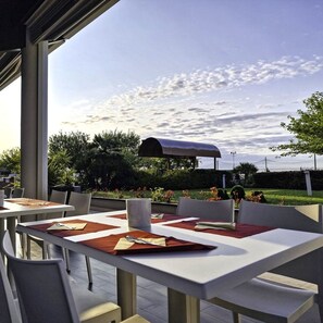 Een terras met tafels en stoelen, een overdekte barbecue en uitzicht op de lucht en begroeiing.