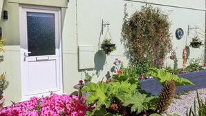 Een gebouw met een witte deur, een klein raam en een aan de muur bevestigd elektriciteitsdoos. Voor het gebouw hangen bloembakken en staan verschillende planten en bloemen.