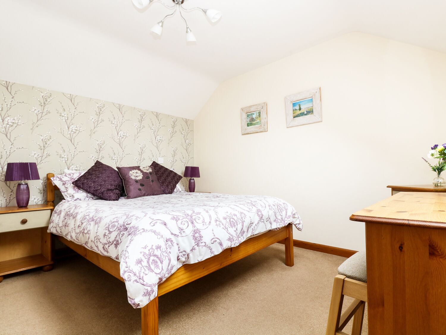 A bedroom with a wooden bed, two nightstands with lamps, a wooden desk, and two framed pictures on the wall.