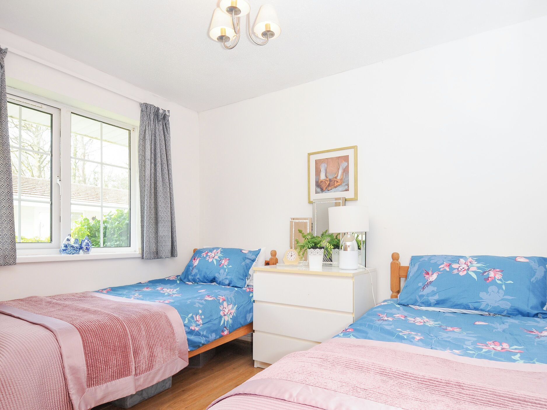 Een slaapkamer met twee bedden, een kaptafel, een raam met gordijnen, een nachtkastje met een lamp en een ingelijst schilderij aan de muur.