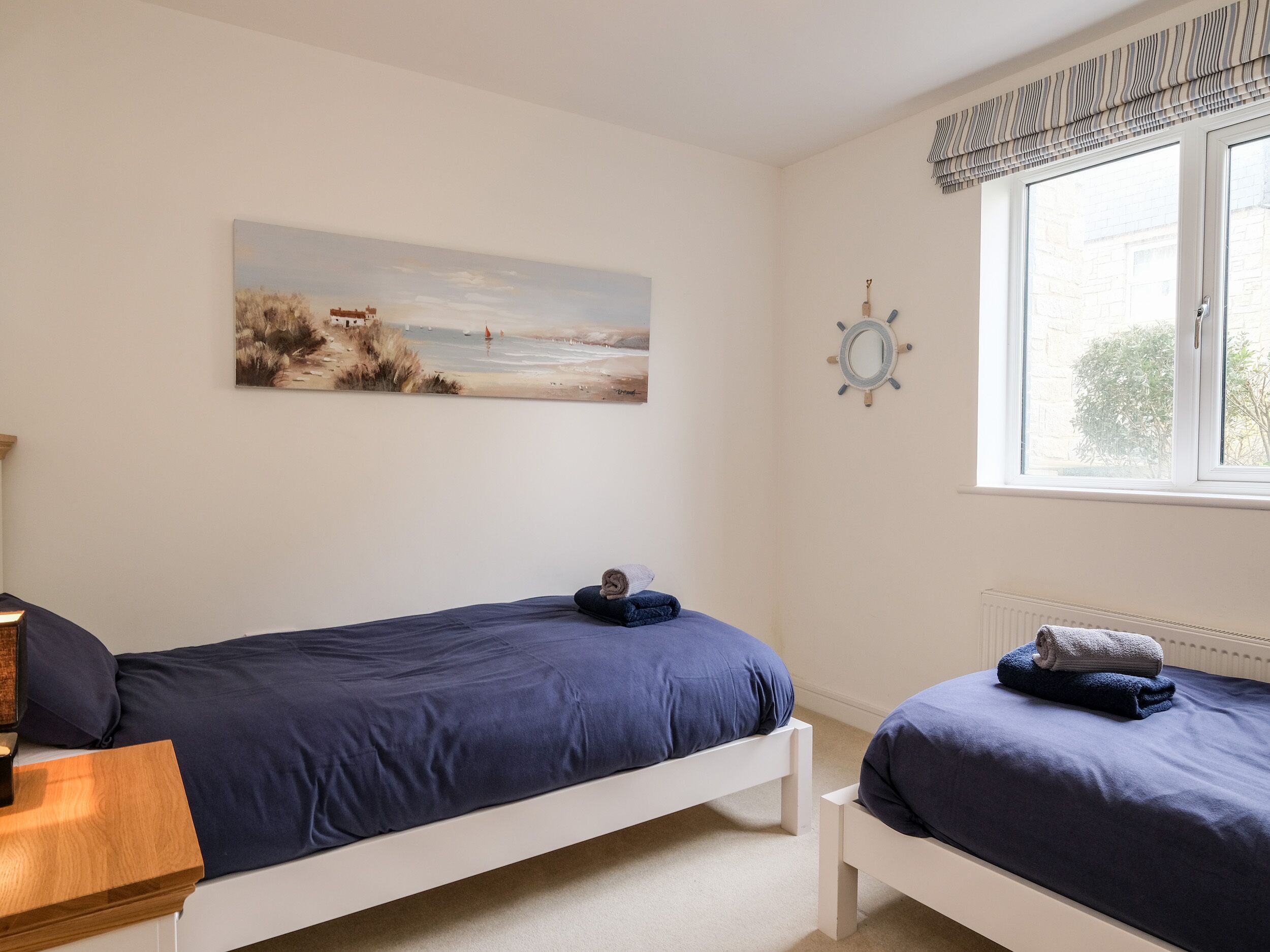 Ein Schlafzimmer mit zwei Einzelbetten, einem Gemälde mit Strandmotiv, einer Schiffslenkrad-Dekoration und einem Fenster mit gestreifter Gardine.