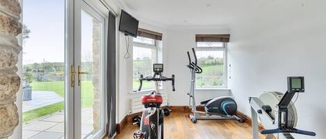 Un gimnasio en casa con bicicleta estática, elíptica y remo, ventanas amplias y una pared de piedra.