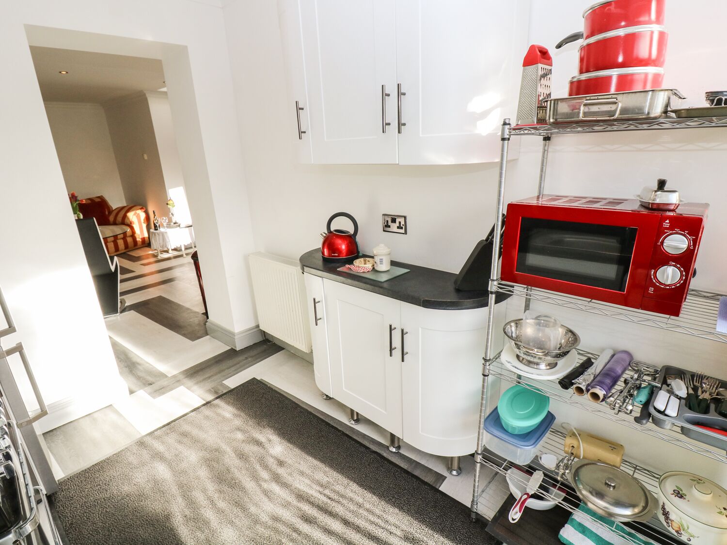 A compact kitchen with white cabinets, a red kettle, a microwave, and a metal rack with kitchen utensils.