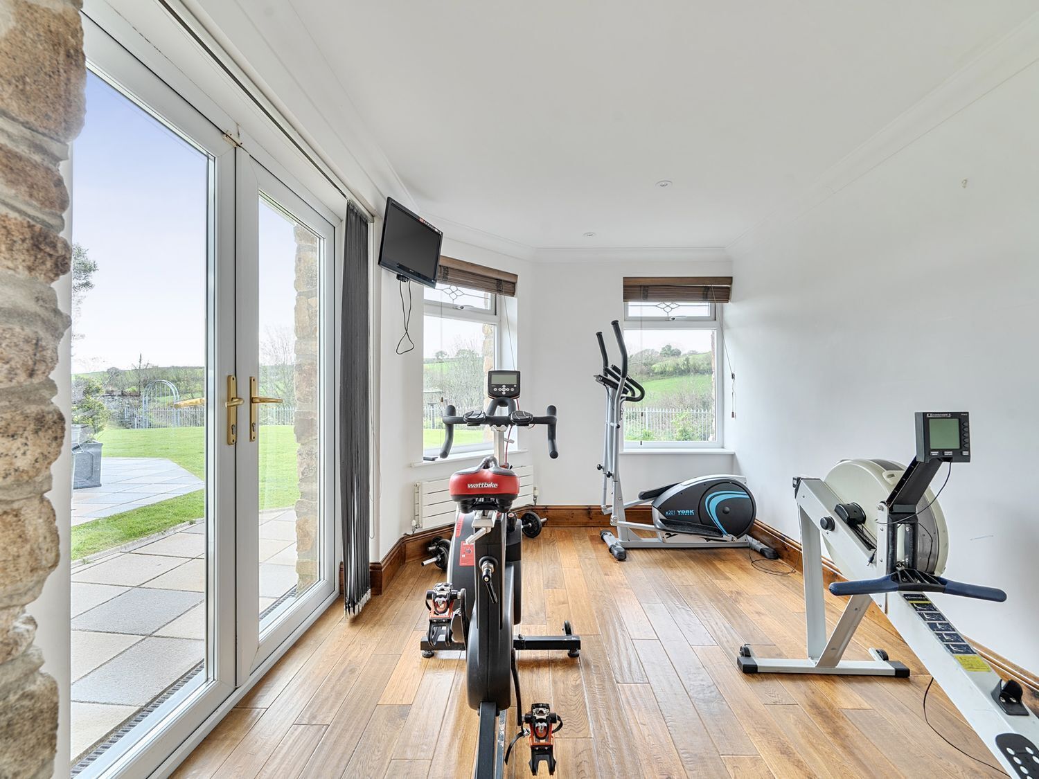 Un gimnasio en casa con bicicleta estática, elíptica y remo, ventanas amplias y una pared de piedra.