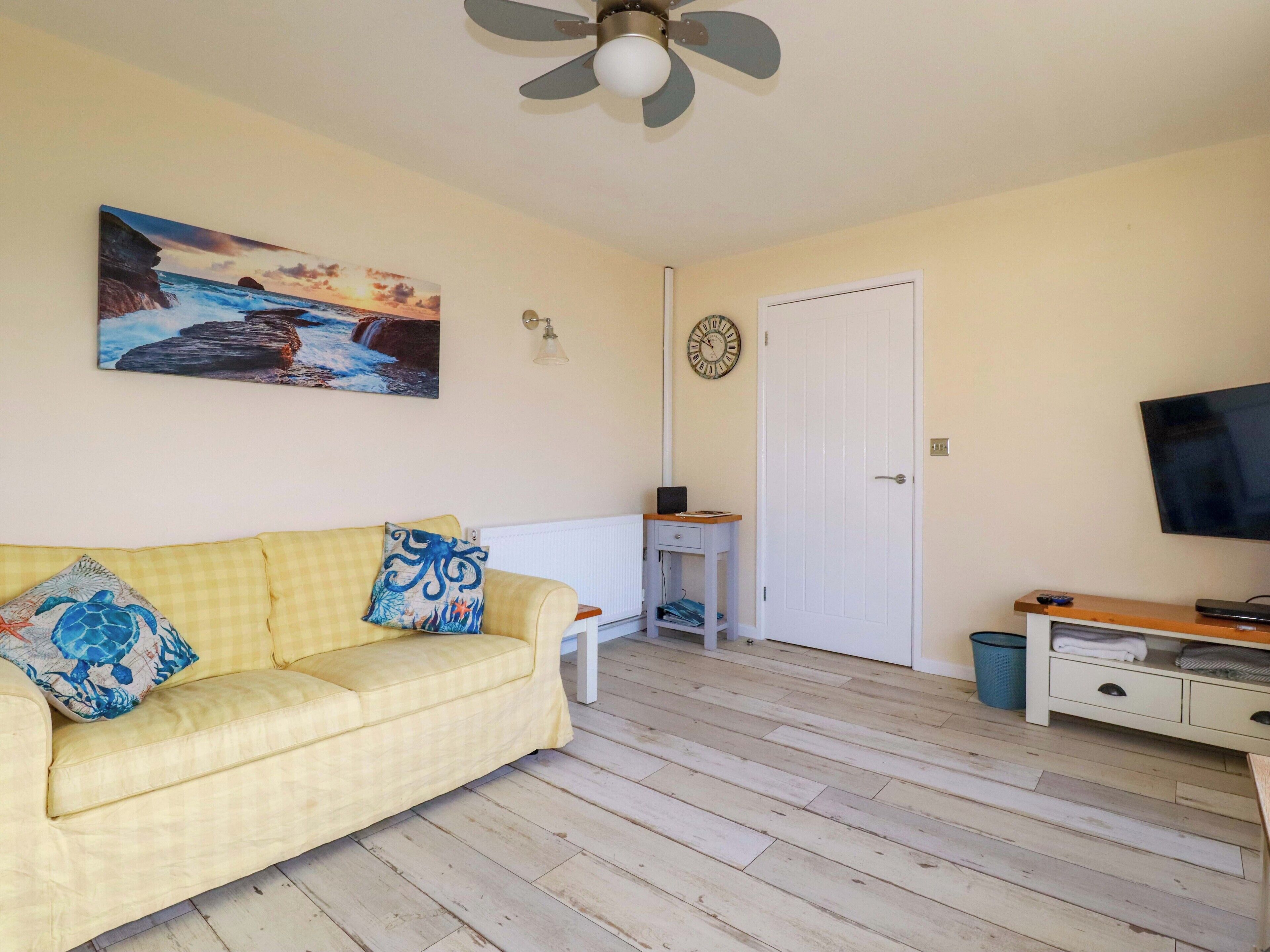 Un salón con sofá amarillo, un cuadro de una playa, ventilador de techo, puerta blanca y televisor sobre un mueble de madera.