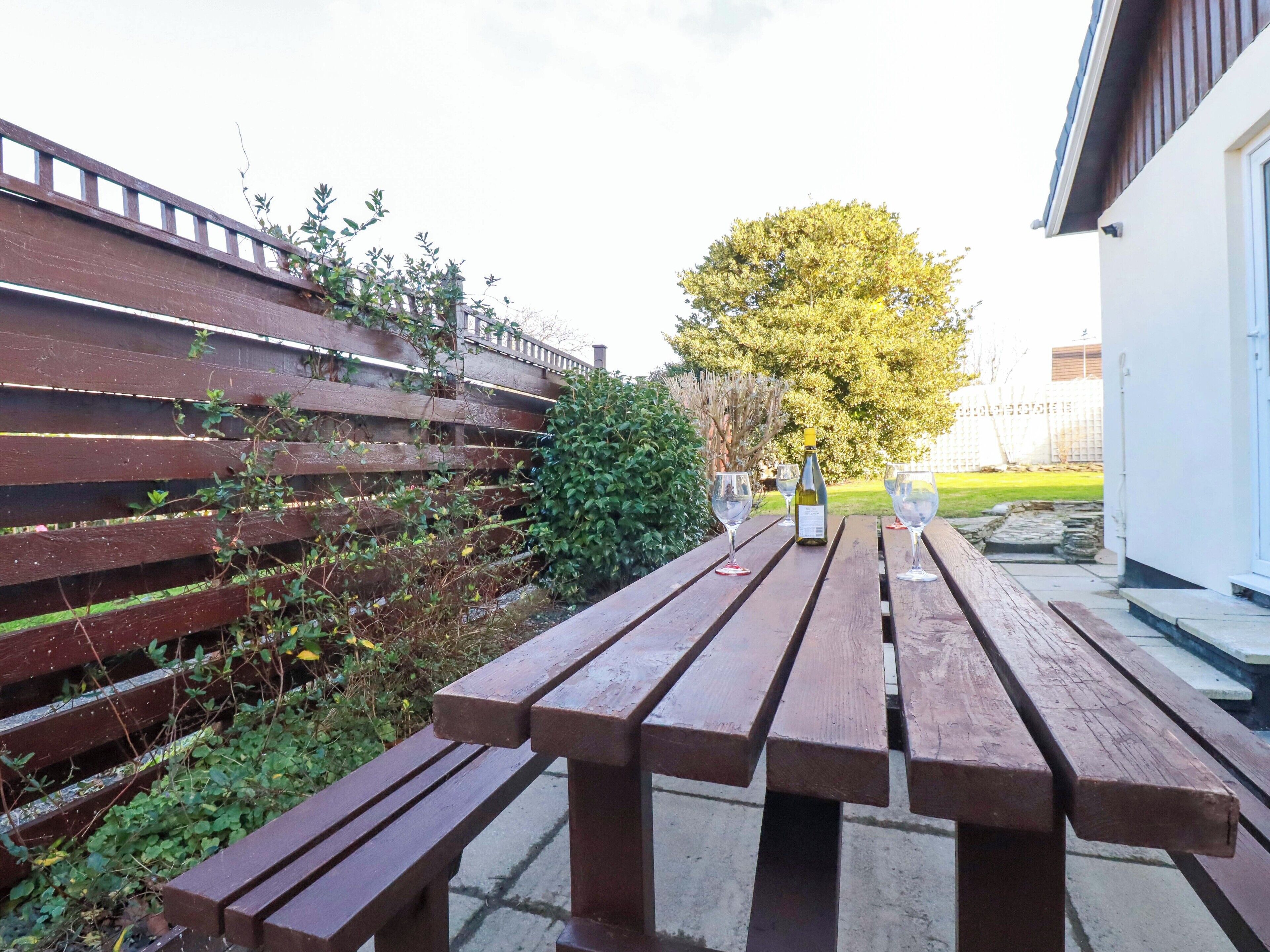 A wooden picnic table with a bottle of wine and glasses, set against a backdrop of a wooden fence and a tree.