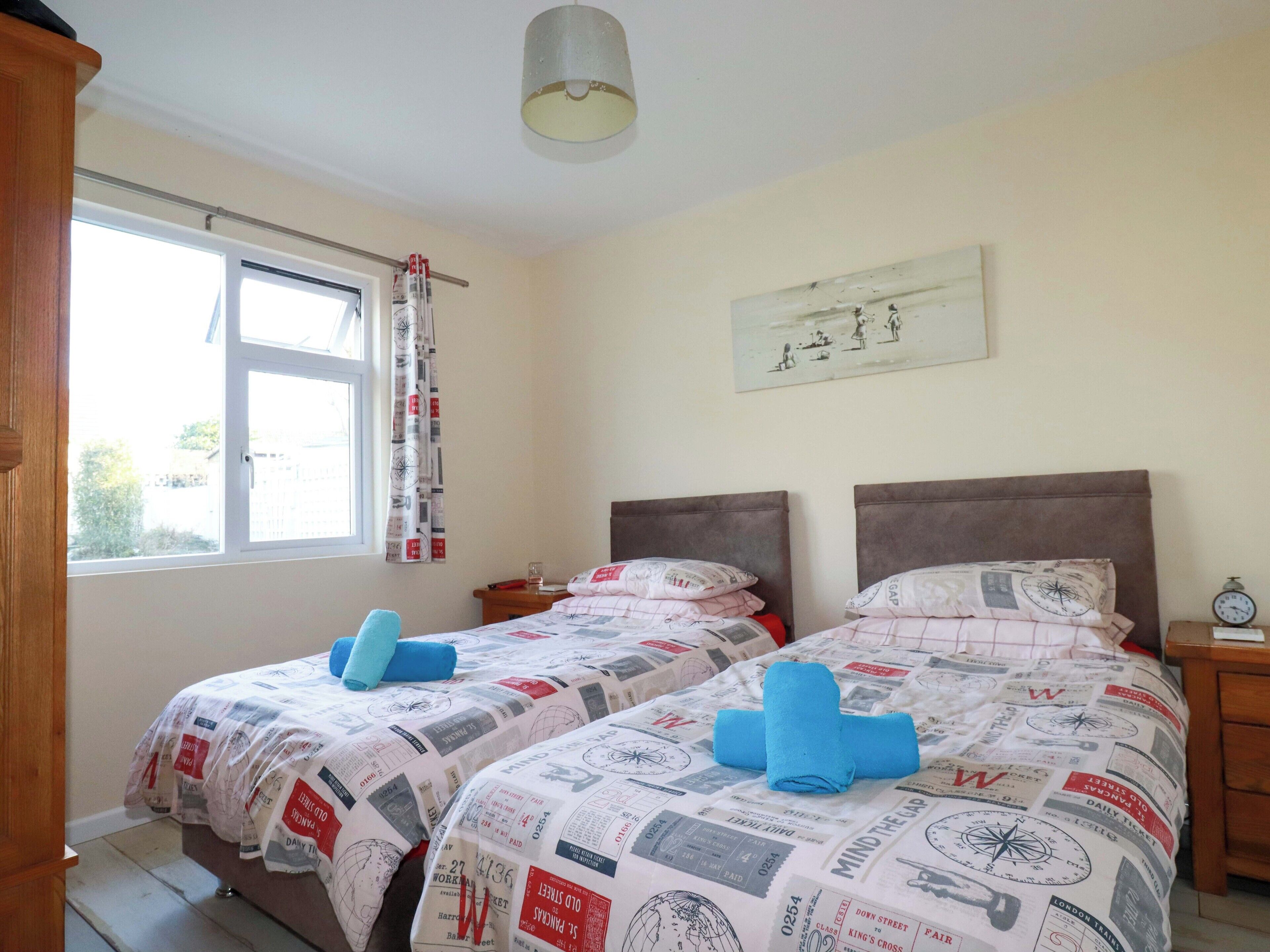 Un dormitorio con dos camas, cada una con una colcha estampada y toallas azules. Hay una ventana con cortinas, una mesita de noche con una lámpara y un reloj de pared.