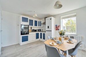 Une cuisine moderne avec des armoires blanches et bleues, un four encastré, un micro-ondes et un réfrigérateur. On y trouve une table à manger avec des chaises et une fenêtre donnant sur la forêt.