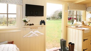 Una habitación acogedora con cama, televisor montado en la pared, ventana con vistas al exterior, una pequeña estantería con un florero y un mueble de madera con un lavabo.
