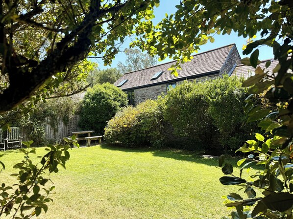 A stone house with a sloped roof, surrounded by a well-maintained lawn and dense shrubbery. - Meadow Cottage (Fowey)