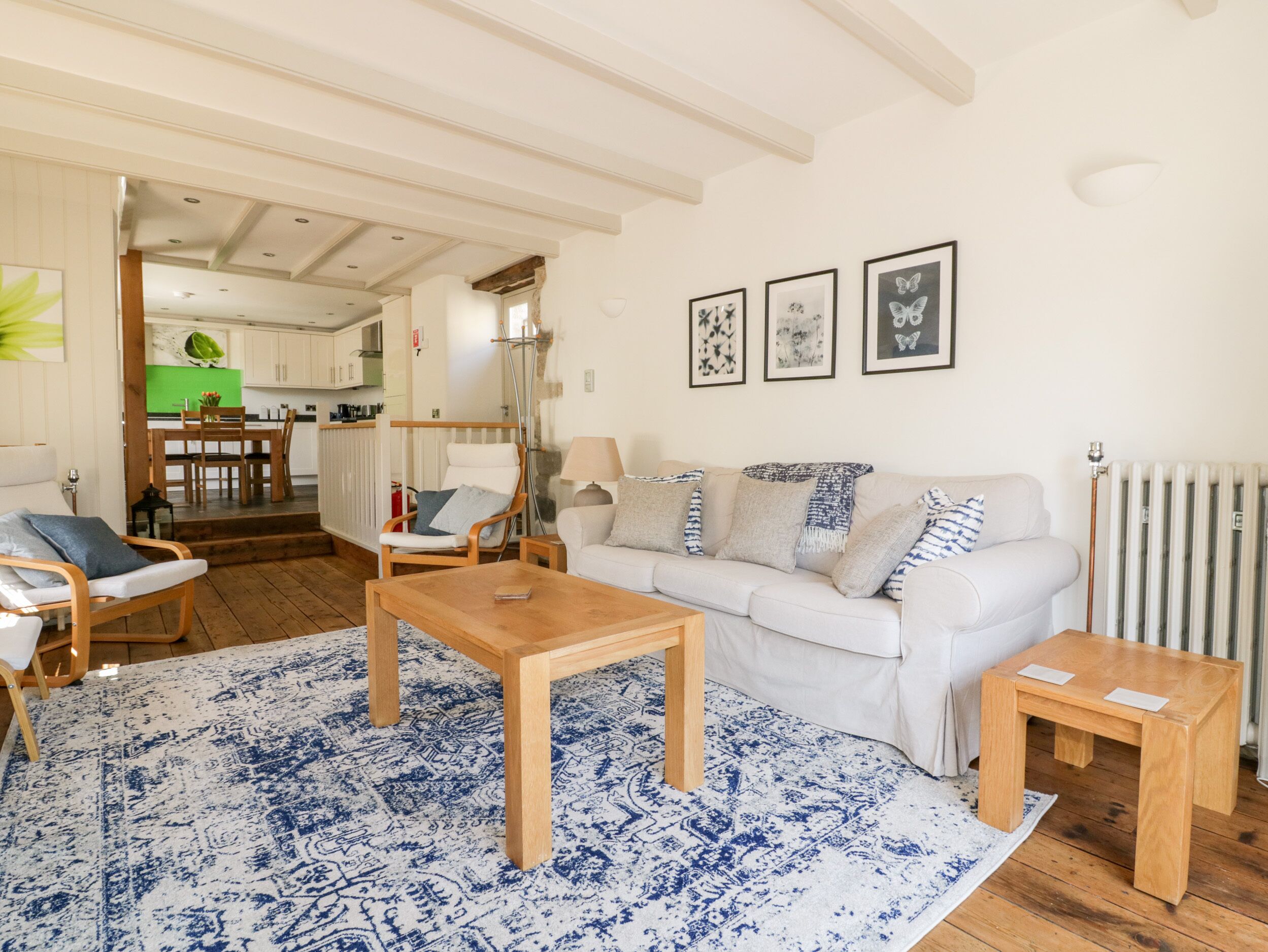 Un salon chaleureux avec un canapé blanc, une table basse en bois et un tapis à motifs bleus. On aperçoit à l’arrière-plan un coin repas avec une table et des chaises vertes.