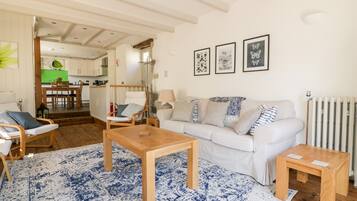 Un salon chaleureux avec un canapé blanc, une table basse en bois et un tapis à motifs bleus. On aperçoit à l’arrière-plan un coin repas avec une table et des chaises vertes.