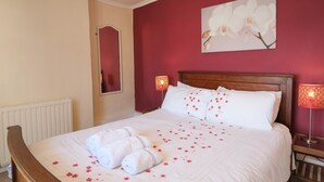 Un lit bien fait, avec du linge de lit blanc et des motifs floraux roses, une tête de lit en bois et un mur rouge orné d’un tableau représentant une orchidée.