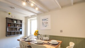 Un coin repas avec une table en bois dressée pour quatre personnes, entourée de chaises blanches. Une étagère contenant divers objets est fixée au mur, sur le côté gauche.