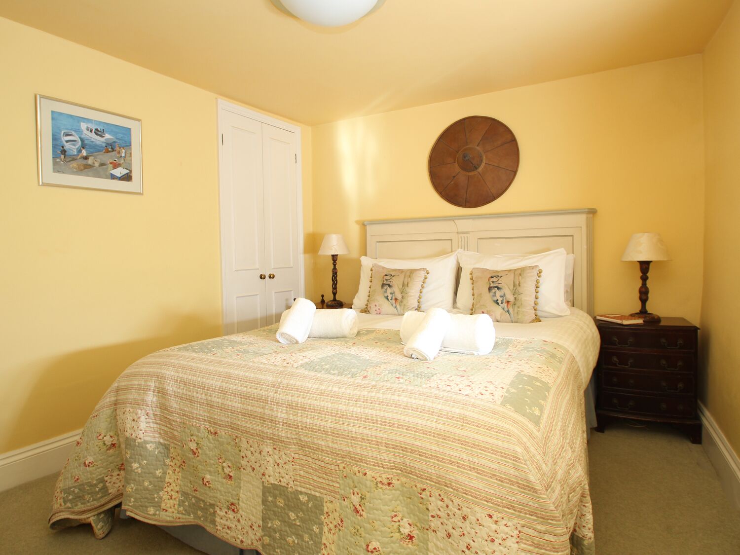A neatly made bed with floral bedding, a round wall clock, and a painting of a boat on the wall.