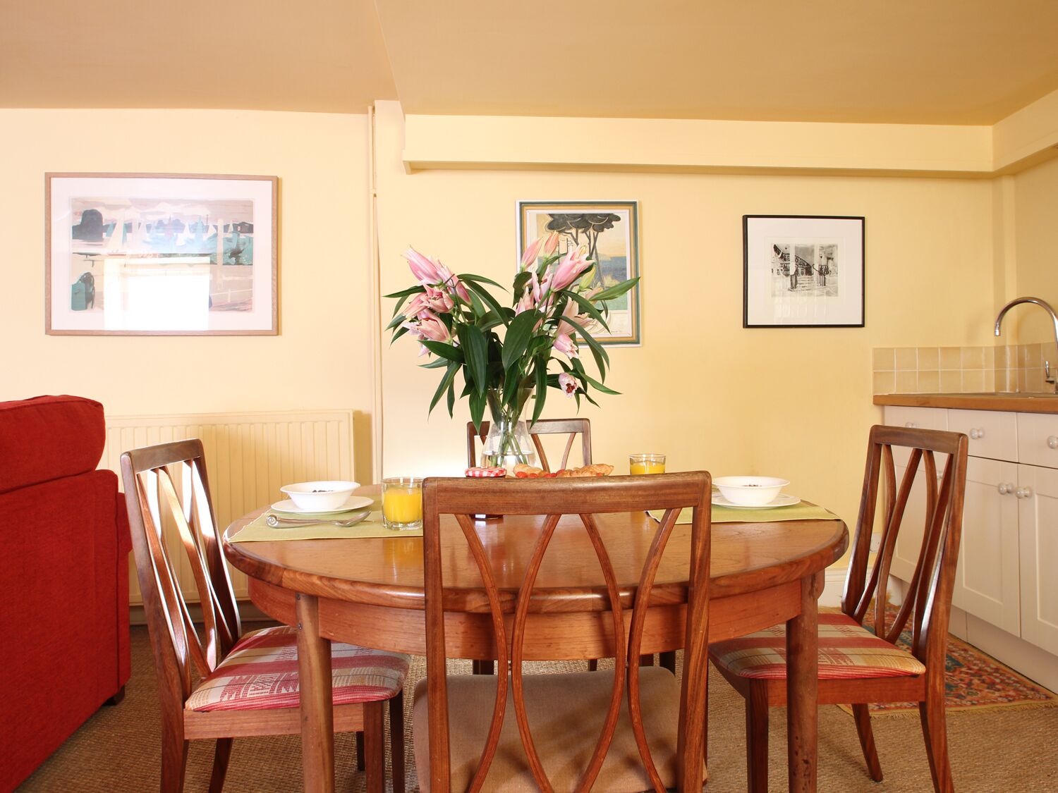 A dining area with a round wooden table, six chairs, a vase of flowers, and framed pictures on the walls.