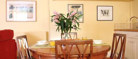 A dining area with a round wooden table, six chairs, a vase of flowers, and framed pictures on the walls.