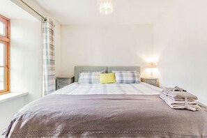 A neatly made bed with a plaid bedspread and pillows, a bedside lamp, and a window with curtains. - Victoria Cottage (Newquay)