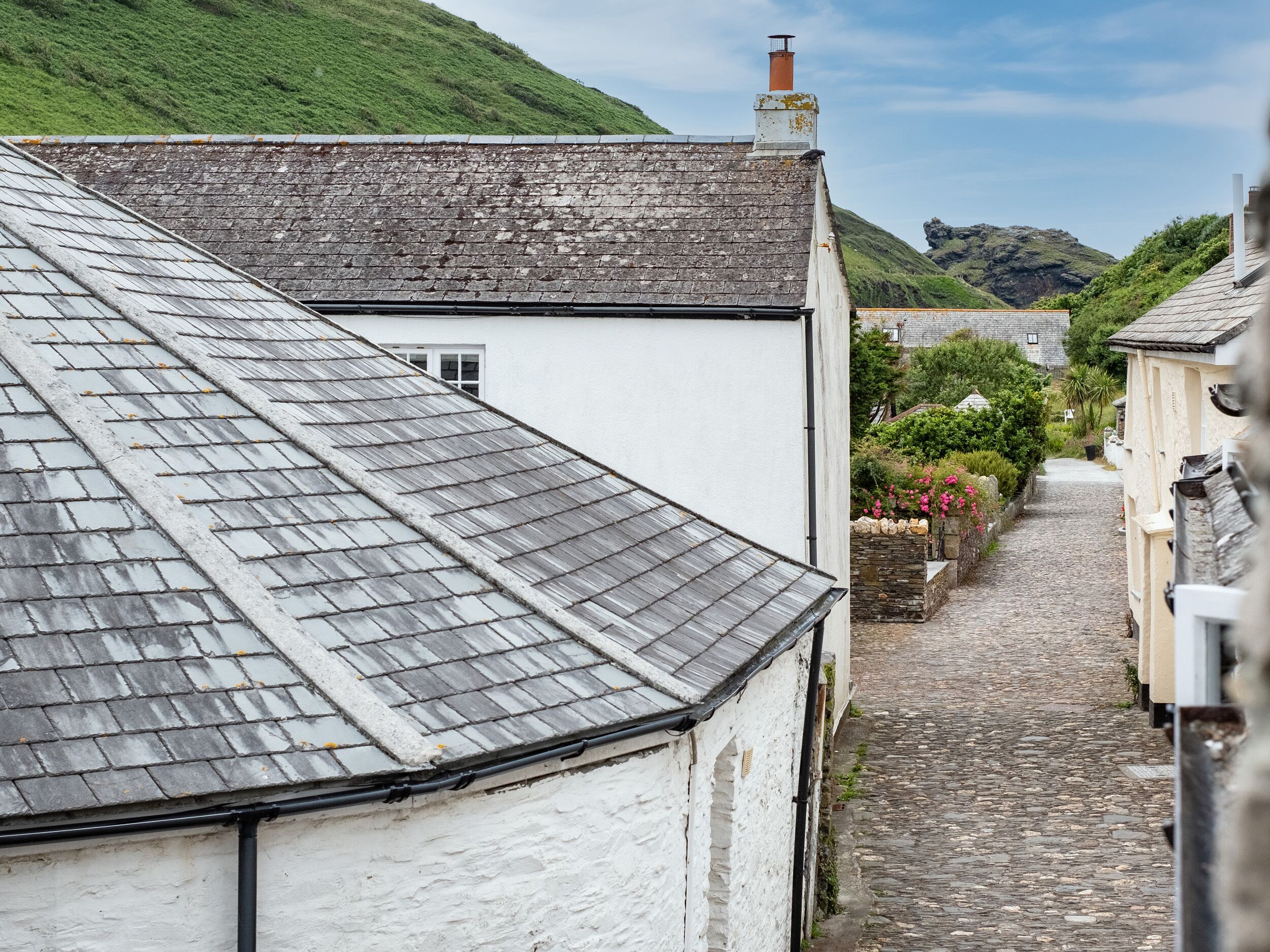 A narrow cobblestone street flanked by traditional white buildings with grey roofs, leading to a hill with a rocky outcrop.
