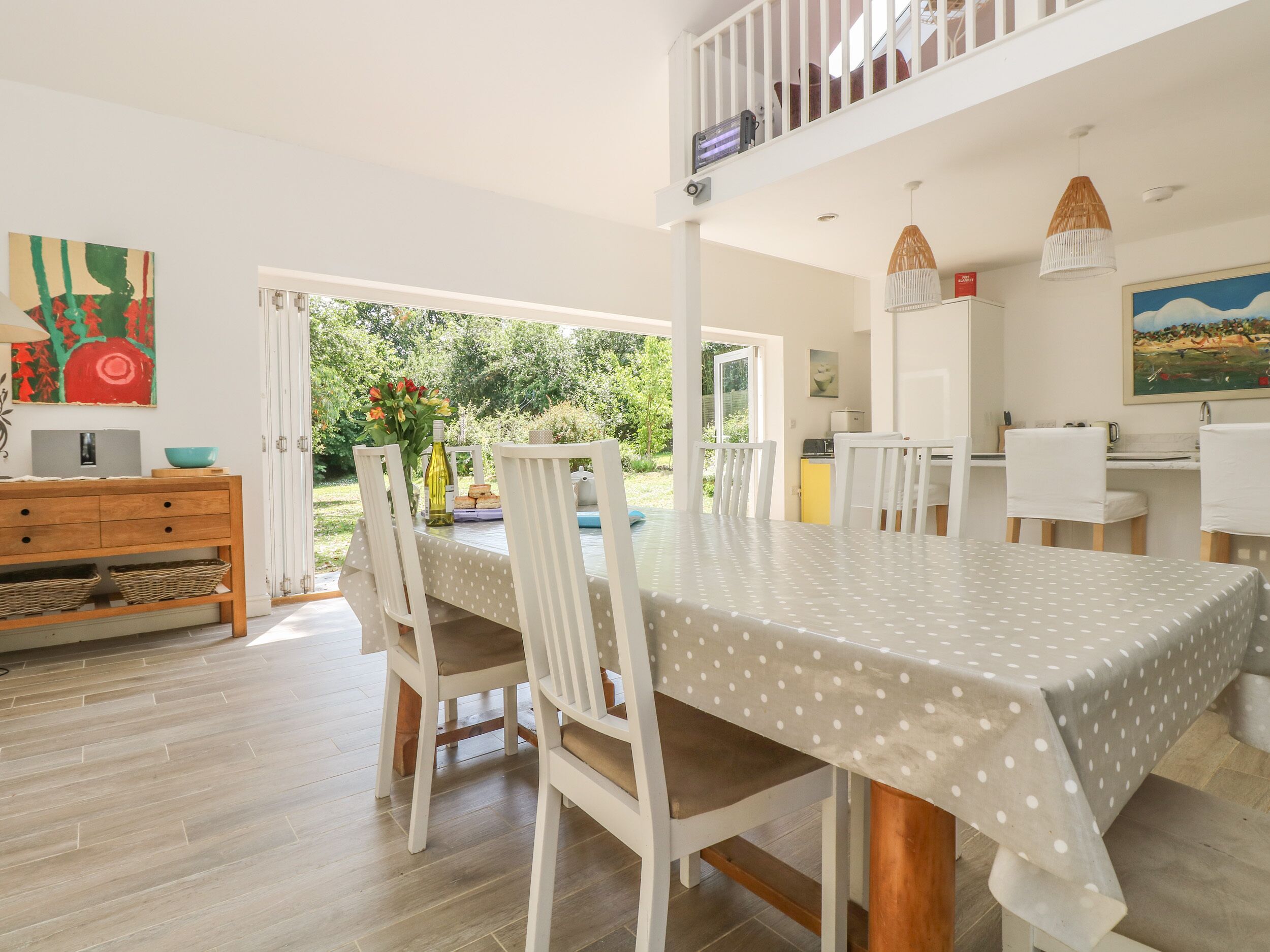 A dining area with a table covered in a polka dot tablecloth, surrounded by white chairs. There is a wooden cabinet with baskets underneath, and a painting on the wall.