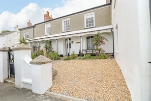 Une maison à deux étages, à l’extérieur blanc, avec une allée en gravier et un petit jardin agrémenté de fleurs roses.