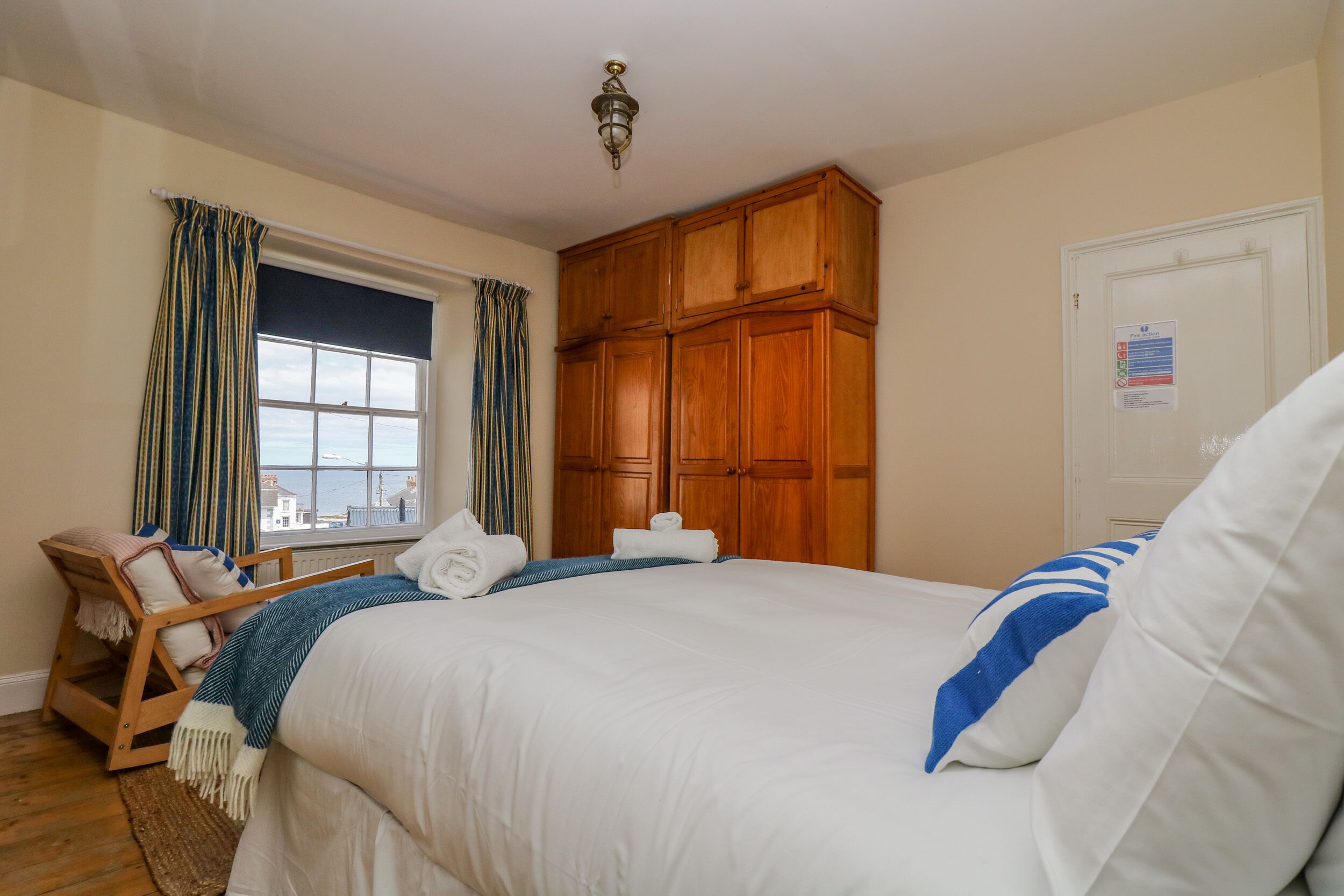 Habitación de hotel con una cama grande, armario de madera, ventana con cortinas, una silla y vistas al mar.