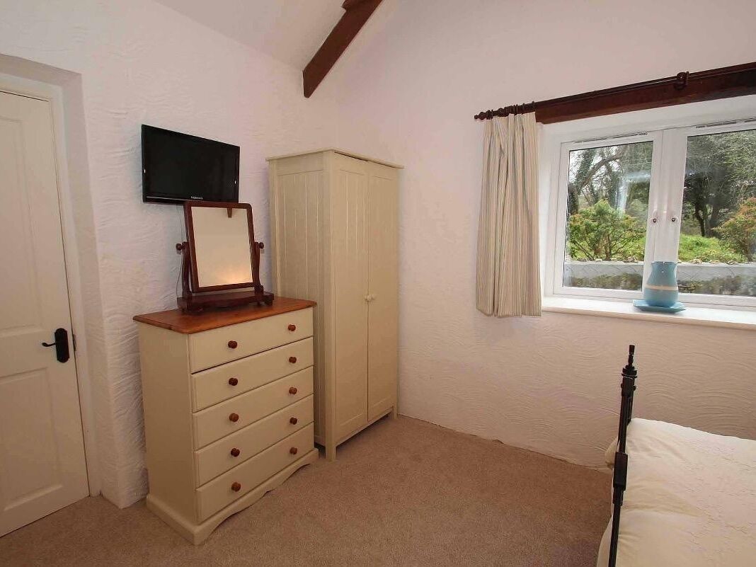 A bedroom with a bed, a dresser, a television, a window with curtains, and a view of greenery outside.