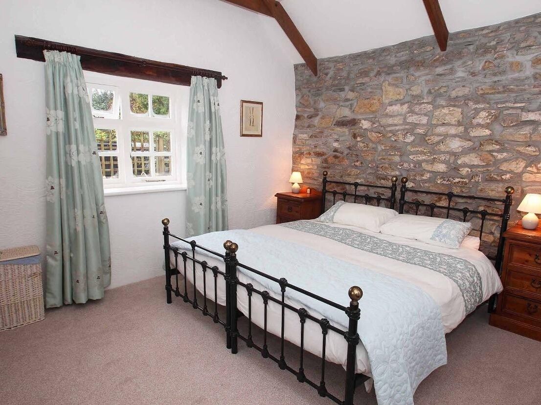 A bedroom with a stone wall, a bed with a metal frame, a window with curtains, and a bedside table with a lamp.