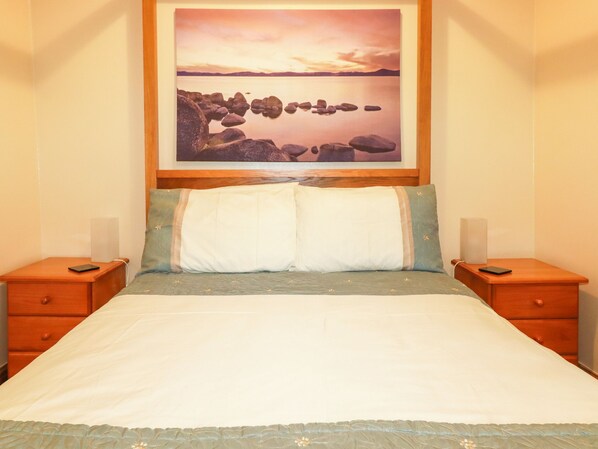 A bed with a white and green bedspread, two pillows, and a framed picture of a sunset over rocks on the wall above. - Barn Cottage (Liskeard)