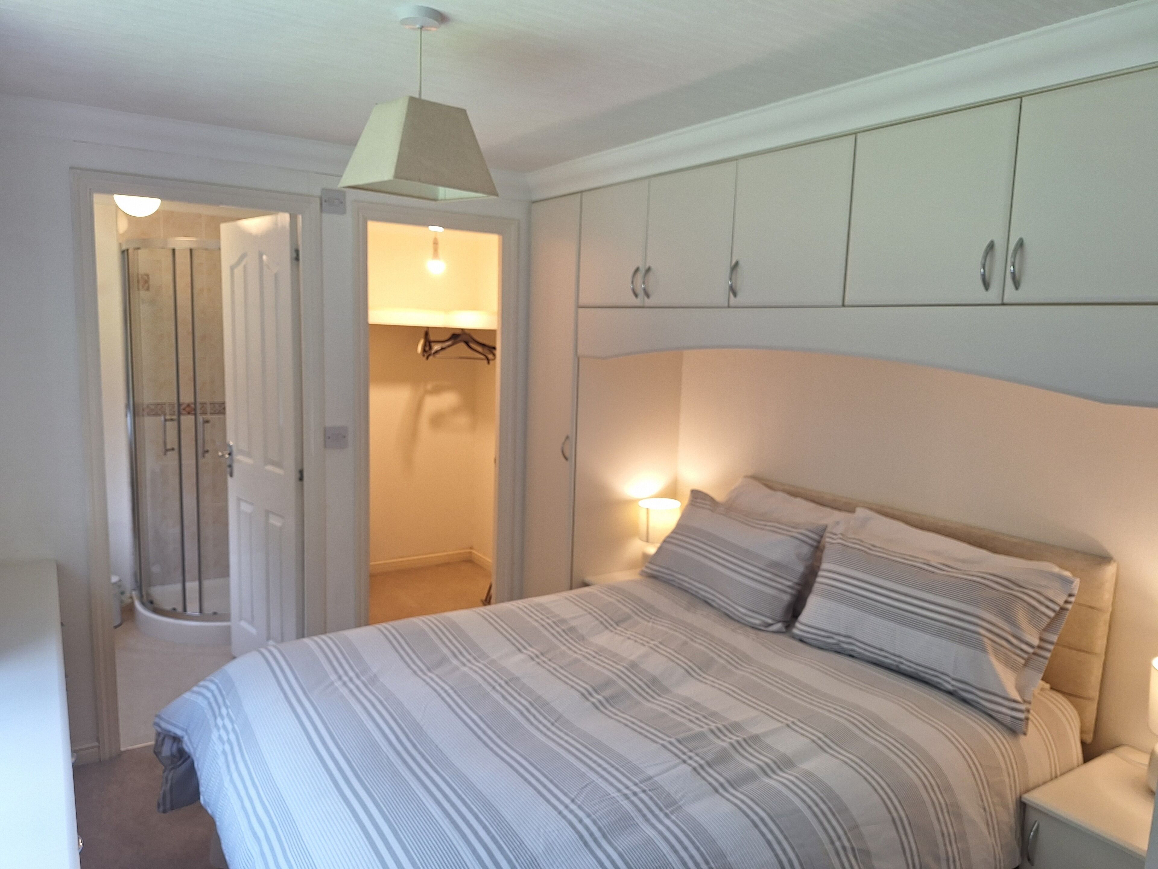 A bedroom with a bed, a nightstand, a lamp, a wardrobe, and a shower area.