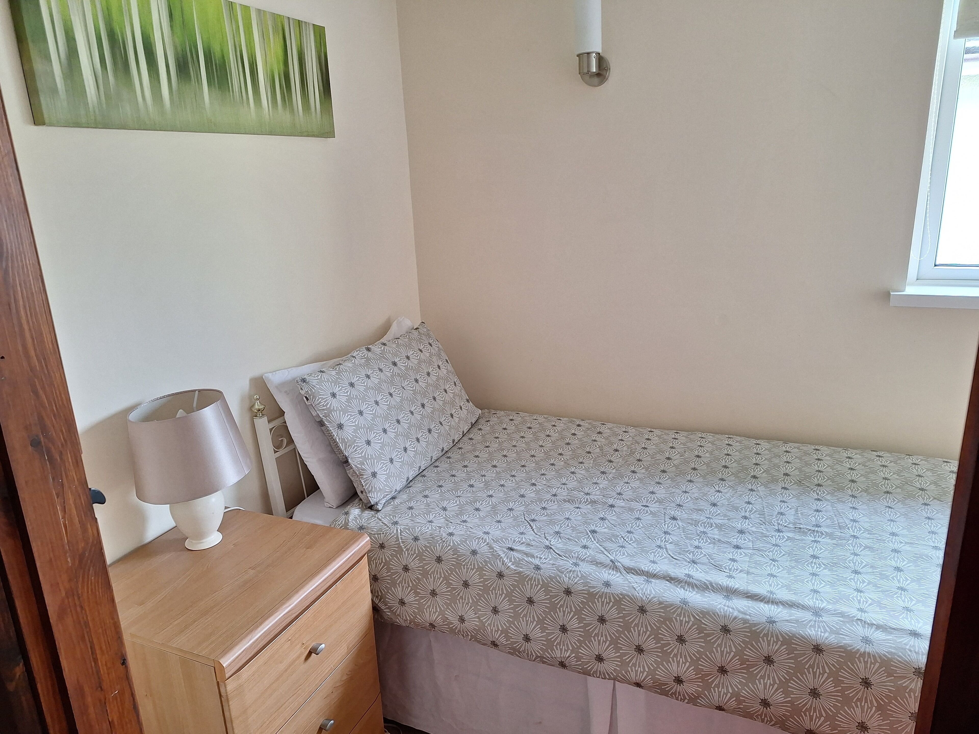 A small, single-bed room with a wooden nightstand, a lamp, and a wall-mounted light fixture.