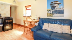 A living room with a blue sofa, wooden dining table, and a kitchen area with an oven. - Cosy Cottage (Liskeard)