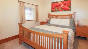 A bedroom with a wooden bed, a nightstand, a bedside table, a window with curtains, and a painting of red flowers on the wall. - Cosy Cottage (Liskeard)