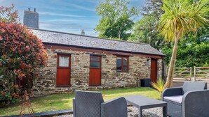 A stone building with a tiled roof, a small porch, and a gravel patio with wicker furniture. - Cosy Cottage (Liskeard)