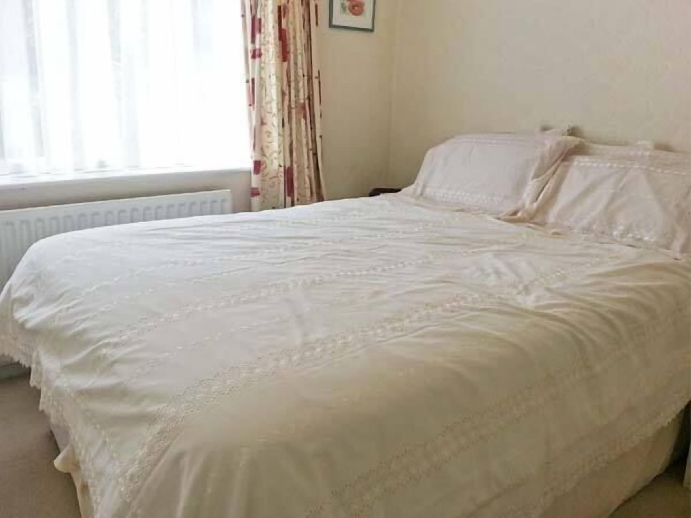 A neatly made bed with white linens in a room with a radiator and a window with curtains.