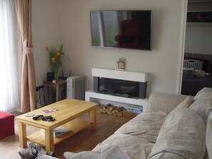 Una sala de estar con una mesa de centro de madera, un sofá, un televisor de pantalla plana montado en la pared, un radiador y una pequeña mesa con un florero.