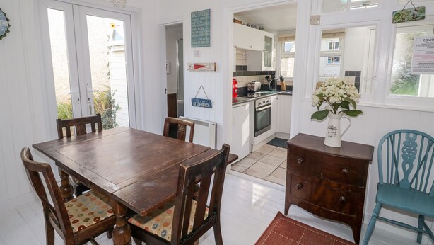 Un coin repas avec une table et des chaises en bois, une cuisine à l’arrière-plan et une vue sur l’extérieur par une porte.
