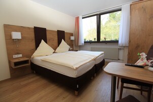 A bedroom with a wooden headboard, a bed with two pillows, a bedside table, a nightstand, a window with curtains, and a wooden desk with a chair.