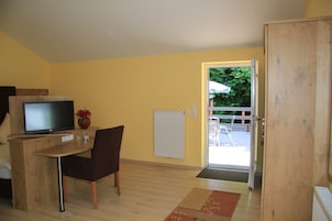 A room with a wooden desk, a television, a chair, a radiator, and a door leading to an outdoor area with a chair and umbrella.